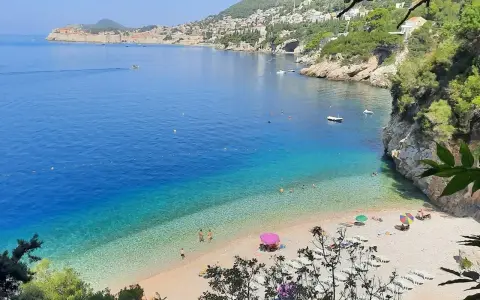 Malownicza zatoka z turkusową wodą i kamienistą plażą. W oddali widać mury Starego Miasta Dubrownik. Idealne plaże Dubrownik na relaks.