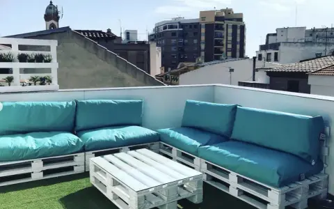 Sofá y mesa de centro hechos con palets blancos, con cojines turquesas, en una terraza urbana. ¡Una idea genial de como hacer un sofa con palets!