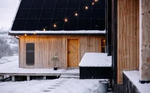Ett modernt solcellsbatteri utomhus monterat på en träfasad, med snö på taket och marken. Julbelysning lyser upp kvällen.