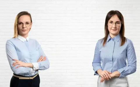 Dwie kobiety w koszulach stoją przed białą ceglaną ścianą.