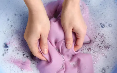 Manos lavando ropa morada a mano en agua con jabón y burbujas.