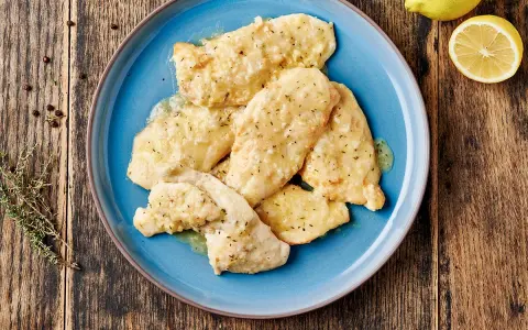 Piatto di pollo al limone su un piatto blu, con limone e rosmarino sullo sfondo.