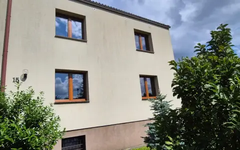 Jasny budynek z oknami o standardowych wymiarach i brązowych ramach, otoczony zielenią.