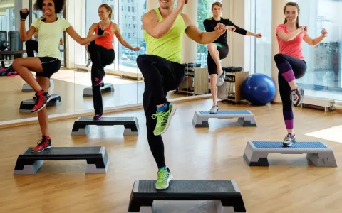 En grupp människor utför step ups i ett gym. De har roligt medan de tränar.