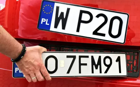 Dłoń z bransoletką montuje tablice rejestracyjne na czerwonym samochodzie. Widać dwie tablice: jedną z napisem WP20, drugą z numerem O-7FM91.