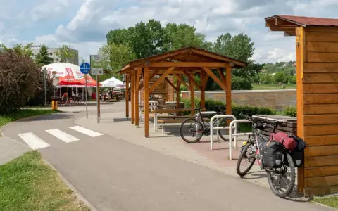 Miejsce odpoczynku przy ścieżce rowerowej. Dwa rowery, w tym jeden z sakwami, czekają na dalszą podróż. W tle zadaszenie i stoliki, idealne na przerwę w trasie.