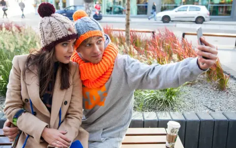 Para do selfie w zimowych stylizacjach. Ona w beżowym płaszczu i czapce z pomponem, on w szarej bluzie i czapce z pomarańczowym akcentem. Jaka czapka do płaszcza? Ta jej wygląda świetnie!