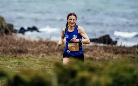 Woman running on a grassy trail by the sea, smiling.
