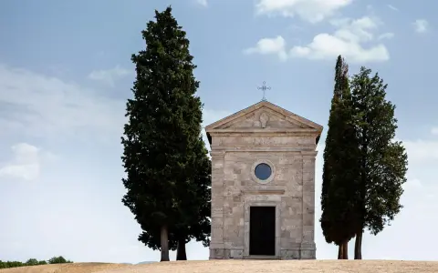 Piccola chiesa in pietra con cipressi, icona del Val d'Orcia, sotto un cielo azzurro con nuvole bianche.