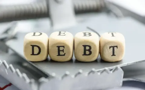 Cztery drewniane kostki z literami D, E, B, T tworzą słowo "DEBT" na pułapce. To symbol finansowych zobowiązań, które mogą prowadzić do trudnej sytuacji, jak datio in solutum.