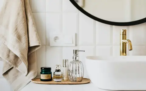 Sur un plateau en bois, des flacons de parfum et un distributeur de savon argenté. Une serviette beige est suspendue à côté d'un lavabo blanc et d'un robinet doré. La colle carrelage flex, c'est quoi ?