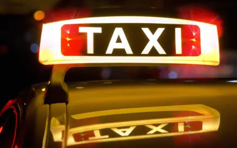 Leuchtendes TAXI-Schild mit roten Blinklichtern, das einen Taxi-Alarm signalisiert. Die Reflexion auf dem dunklen Auto verstärkt die nächtliche Atmosphäre.
