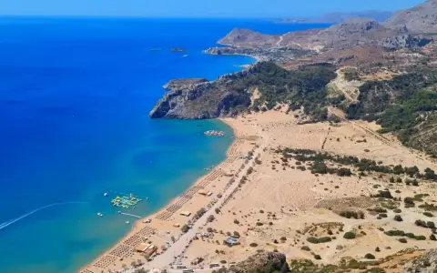 Malownicze **Rodos plaże** z turkusową wodą, piaszczystym brzegiem i zielonymi wzgórzami w tle. Widać wodny plac zabaw.