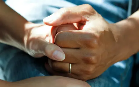 Två händer håller varandra, en gest som visar medlidande och stöd. En tunn ring syns på ena handen.