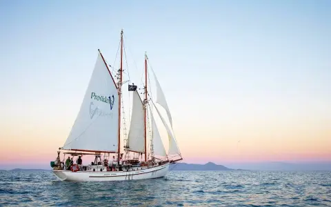 Żaglowiec "Providence" płynie po spokojnych wodach, zbliżając się do Whitehaven Beach. Zachód słońca maluje niebo pastelowymi barwami.
