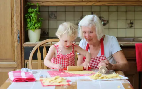 Babcia z wnuczką pieką ciasteczka. Dziewczynka wałkuje ciasto, a babcia z uśmiechem patrzy na jej pracę.