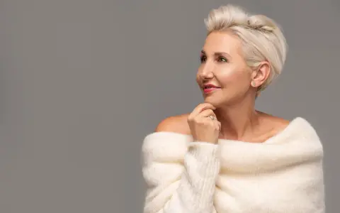 Le visage d'une femme de 80 ans, aux cheveux blonds courts, porte un pull blanc en maille. Elle regarde sur le côté, la main sous le menton.