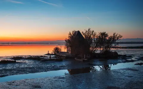Tramonto sulla spiaggia di Marano, con una piccola capanna e un'imbarcazione riflessi sull'acqua calma.