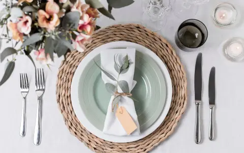 Une table dressée avec une assiette verte, une serviette blanche décorée d'une branche d'eucalyptus et d'une étiquette, le tout sur un rond de serviette fait maison.