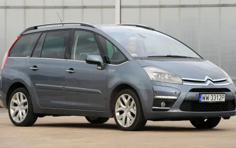 Szary Citroen Picasso z napisem "Picasso" na błotniku, zaparkowany przed budynkiem.