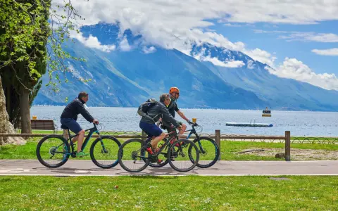 Ciclisti godono di una giornata soleggiata lungo uno dei laghi nord Italia, con montagne maestose sullo sfondo.