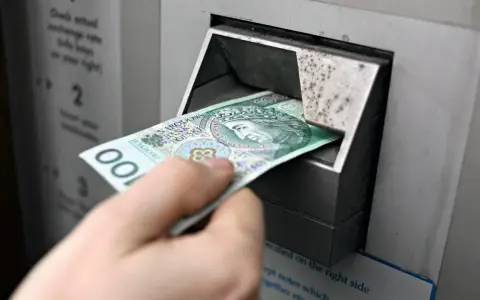 Ręka wkłada banknot 100 zł do bankomatu. Czy państwo może zabrać pieniądze z konta w razie wojny?