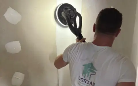 Un ouvrier utilise une ponceuse girafe pour poncer un mur en plâtre, préparant la surface pour la peinture.