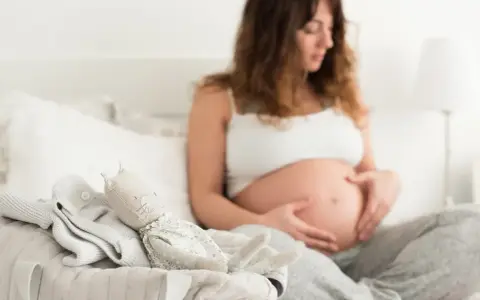 Czekając na pierwsze kopnięcia, przyszła mama głaszcze brzuszek. Obok leżą ubranka i przytulanka dla maluszka.