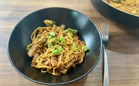 Pyszny makaron wok z kawałkami wołowiny i brokułami, posypany szczypiorkiem. Danie prezentuje się apetycznie w czarnej misce.