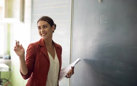 Lehrerin mit Kreide und Unterlagen vor einer Tafel mit einer Matheaufgabe. Sie lächelt und wirkt engagiert, was auf einen interaktiven Unterricht hindeutet.