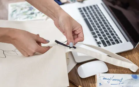 Ręce tworzą torbę DIY, przyszywając czarną wstążkę do płóciennego materiału. Obok leży laptop i myszka.