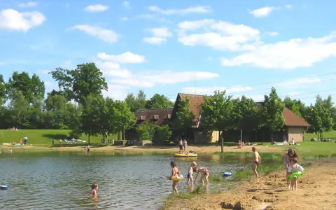Sonniger Tag am badesee fredenbeck. Kinder planschen im Wasser und am Sandstrand, während im Hintergrund Gebäude und Bäume zu sehen sind.