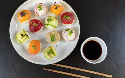 Na talerzu ułożone są kolorowe kulki sushi z łososiem, wołowiną, awokado i rzodkiewką. Obok miseczka sosu sojowego i pałeczki.