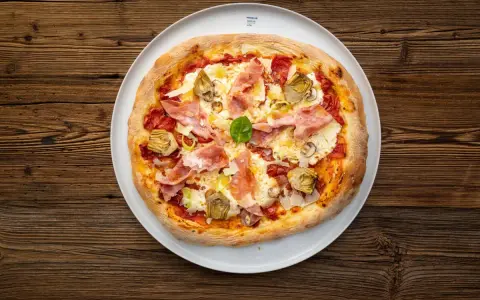 Eine köstliche Prosciutto Pizza mit Artischocken und Basilikum auf einem weißen Teller, serviert auf rustikalem Holz.