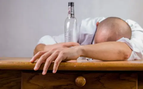 Mężczyzna w białej koszuli leży na stole, głowa oparta na ramieniu. Obok pusta butelka, symbolizująca skutki spożycia alkoholu, jakimi mogą być wymioty po alkoholu.
