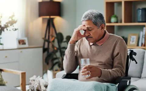 Älterer Mann im Rollstuhl mit Kopfschmerzen, hält ein Glas Wasser. Möglicherweise ein Parkinson-Test der Haut, der auf seine Symptome hinweist.