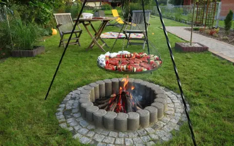 Ognisko w kamiennym kręgu, nad którym wisi okrągła taca z mięsem i warzywami na szaszłykach, gotowe do grillowania w ogrodzie.