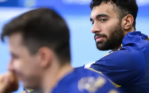 Hassan Kaddah, z brodą i kręconymi włosami, w niebieskiej bluzie z żółtymi detalami, spogląda w bok.
