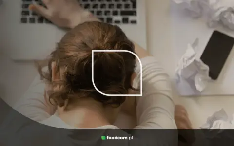 Zmęczona osoba z głową na biurku, otoczona pogniecionymi kartkami i telefonem. To obraz stresu i wypalenia, które mogą być oznaką faz pracoholizmu.