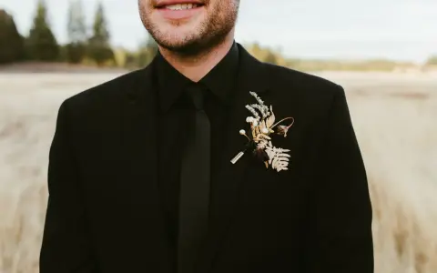 A groom in all black wedding guest attire men's suit, complete with a black tie and a rustic boutonniere.