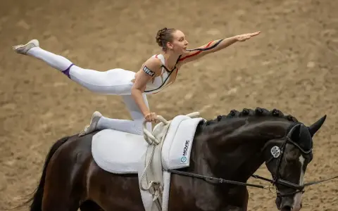Eine Voltigiererin in weißem Anzug balanciert auf einem dunklen Pferd, bereit für das Voltigiieren Olympia.