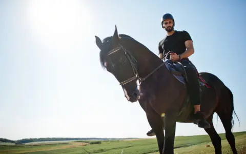 Uomo con barba e casco cavalca un cavallo scuro in un paesaggio verdeggiante, un'esperienza di **Toscana a cavallo**.