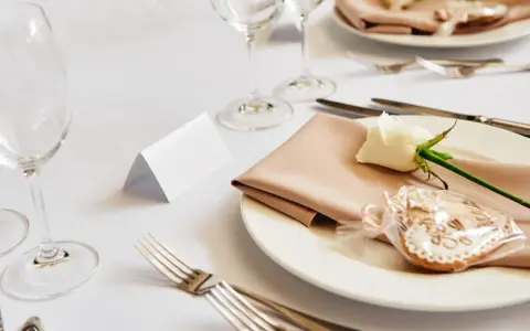 Elegancko nakryty stół weselny z różową serwetką, białą różą i pierniczkiem. Idealne miejsce na zastanowienie, jakie wino na wesele wybrać.