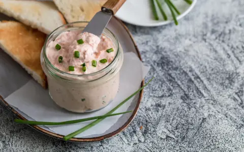 Un pot de rillettes de saumon crémeuses, garni de ciboule, prêt à être tartiné sur des toasts grillés.