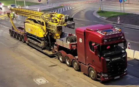 Czerwona ciężarówka Scania z przyczepą transportuje żółtą maszynę wiertniczą. Pilot pojazdu nienormatywnego czeka na przejazd.