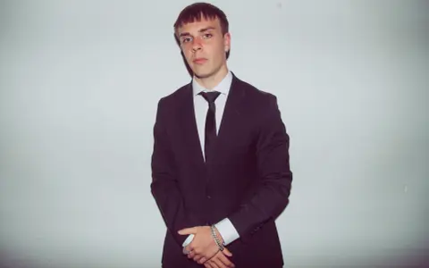 Młody mężczyzna w czarnym garniturze i białej koszuli, z krawatem. Kinny Zimner prezentuje się elegancko.