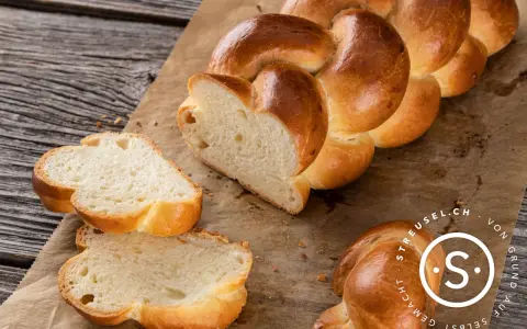 Ein goldbraunes Zopf Brot, frisch gebacken, liegt auf Backpapier. Zwei Scheiben sind abgetrennt und zeigen die weiche Krume.