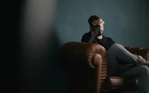 Uomo seduto su un divano di pelle, con la mano sul viso, immerso in pensieri di rimpianto e rimorso.