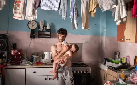 Emiliano Dante, padre single, nutre il suo bambino in una cucina disordinata, con panni stesi ad asciugare.