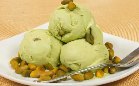 Tre palline di cremoso gelato al pistacchio, guarnite con pistacchi interi, su un piatto bianco con una spolverata di pistacchi tostati.